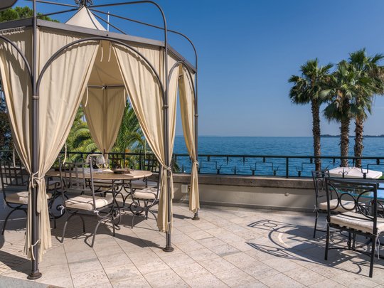A laid-back holiday on Lake Garda at a 4-star hotel Seating area with gazebo overlooking lake and palm trees on a sunny day