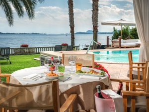 A laid-back holiday on Lake Garda at a 4-star hotel Table with food and wine by pool and lake with palm trees
