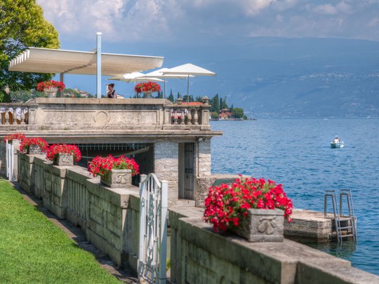A laid-back holiday on Lake Garda at a 4-star hotel Terrace with red flowers by lake with mountains and boat in the background
