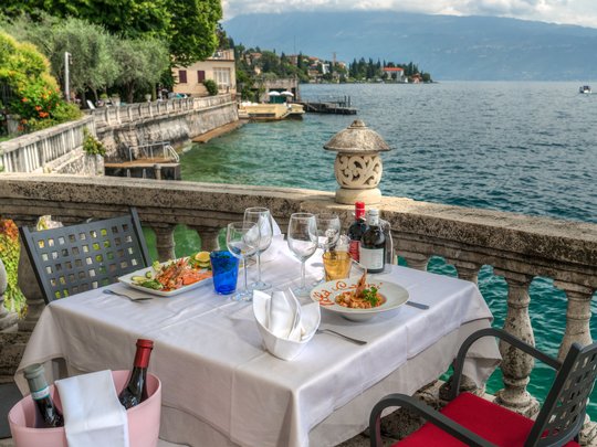 A laid-back holiday on Lake Garda at a 4-star hotel Table with food and wine on a balcony overlooking a lake and mountains